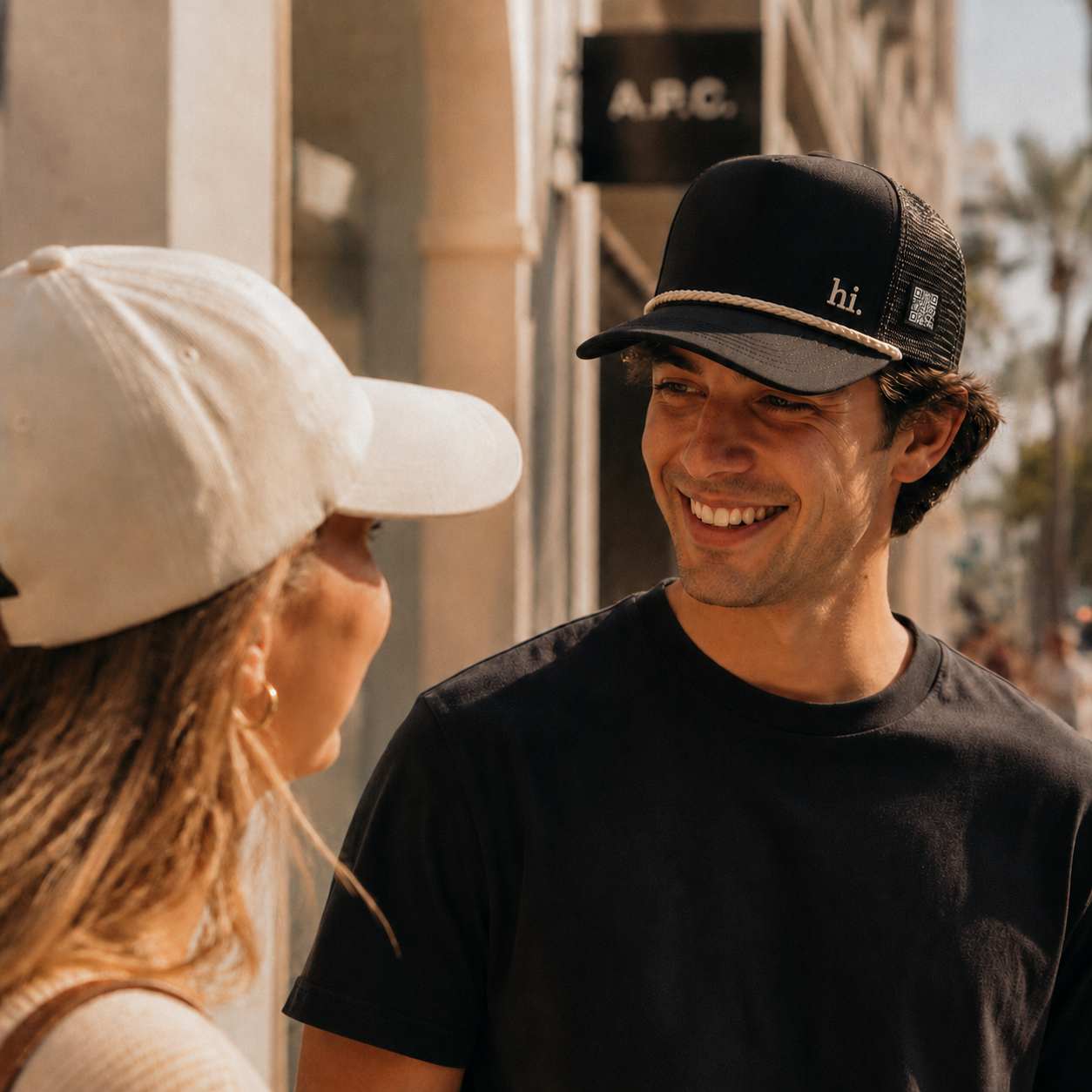 A young couple smiling at each other on a sunny sidewalk — he's wearing the uSayHi black rope trucker hat with a small cream 'hi.' embroidered on the front